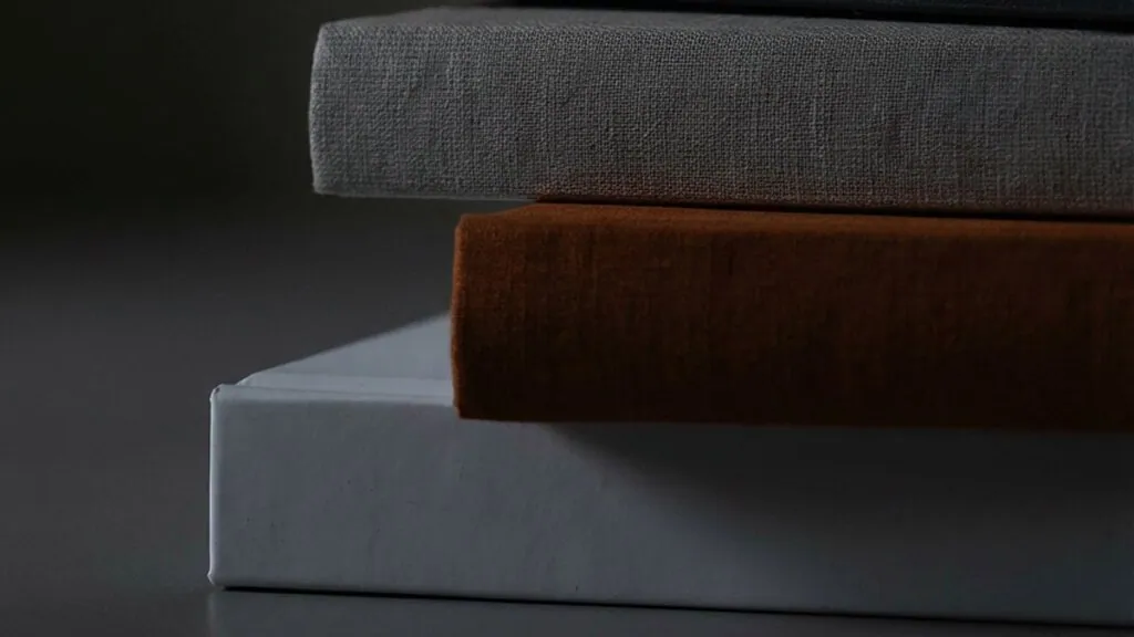 Primer plano de tres libros apilados con lomos de tela en gris, marrón y blanco liso.